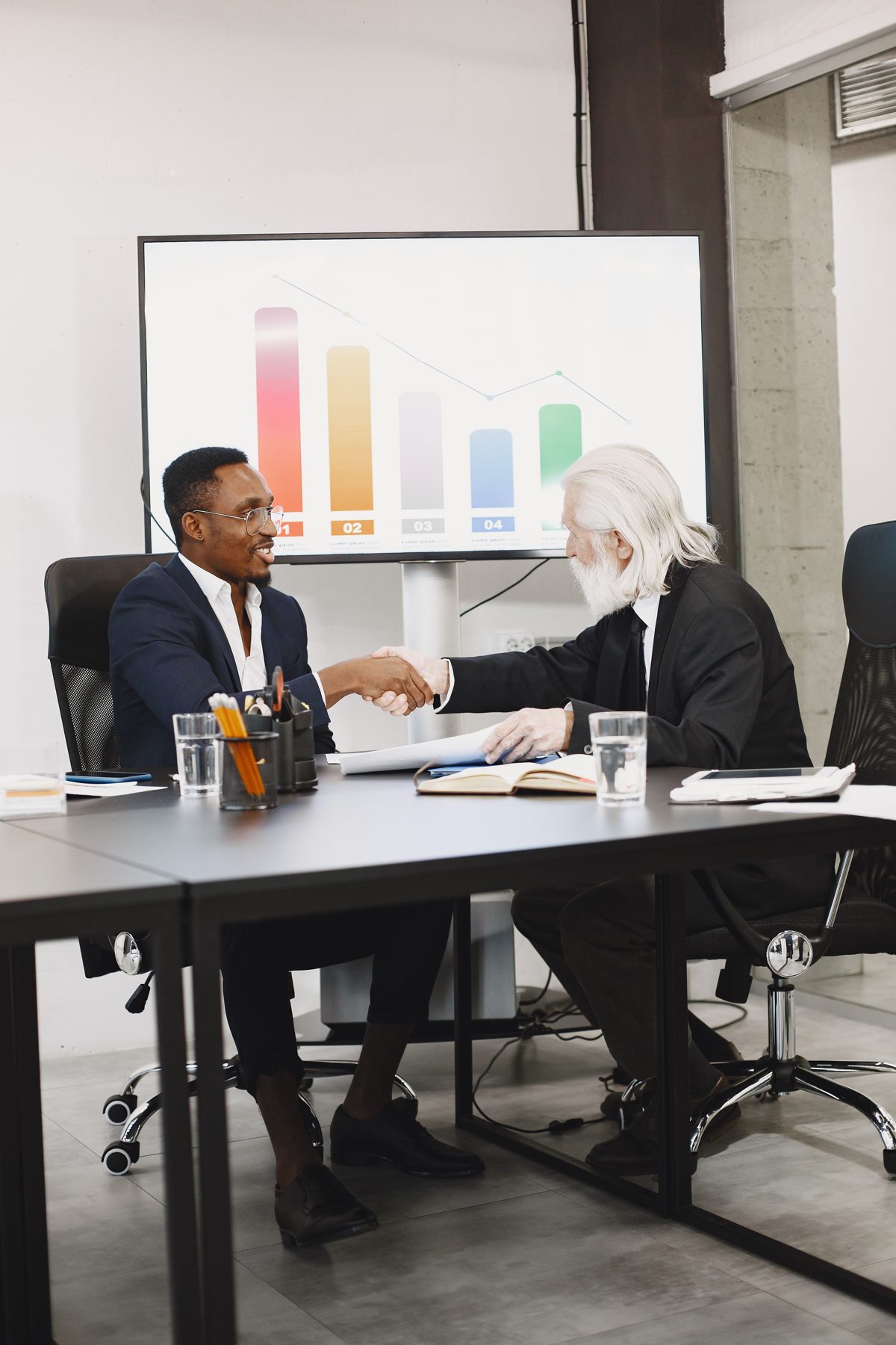 Two advisers shaking hands across a meeting table, with a chart on the screen behind them