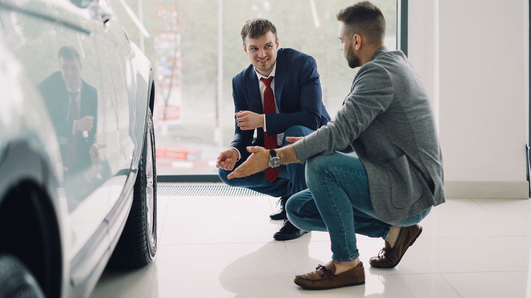 Two men crouched beside a car, talking in the showroom