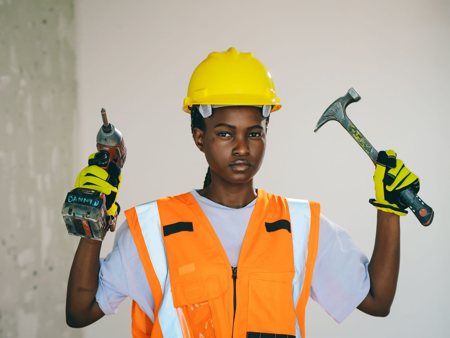 A smiling electrician in a yellow hard hat holding a drill and a hammer